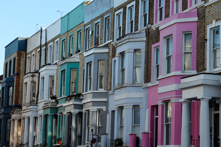 A row of terraced houses with colorful facades in pastel shades of blue, teal, green, yellow, white, and pink, featuring bay windows and decorative mouldings. The buildings are three to four stories t